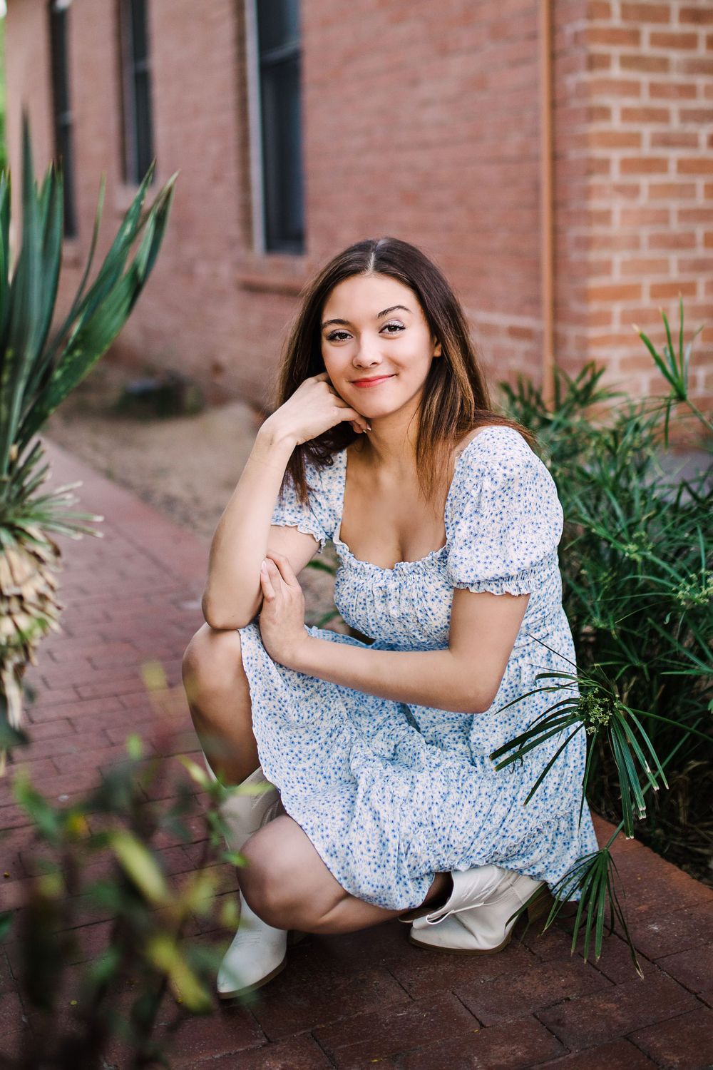 Young woman in blue floral dress crouching on brick path surrounded by green plants near brick wall.
