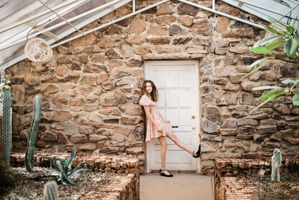 Young woman in floral dress playfully posing by white door in stone greenhouse with cacti.