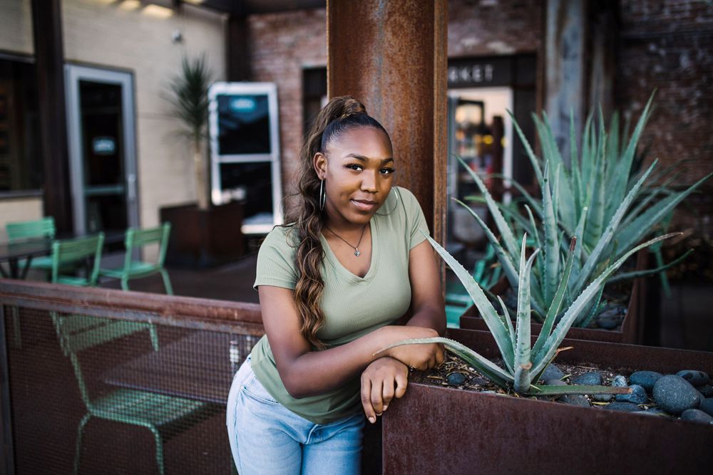Young Black woman in olive tee and jeans leans on rustic metal planter with agave at outdoor patio.