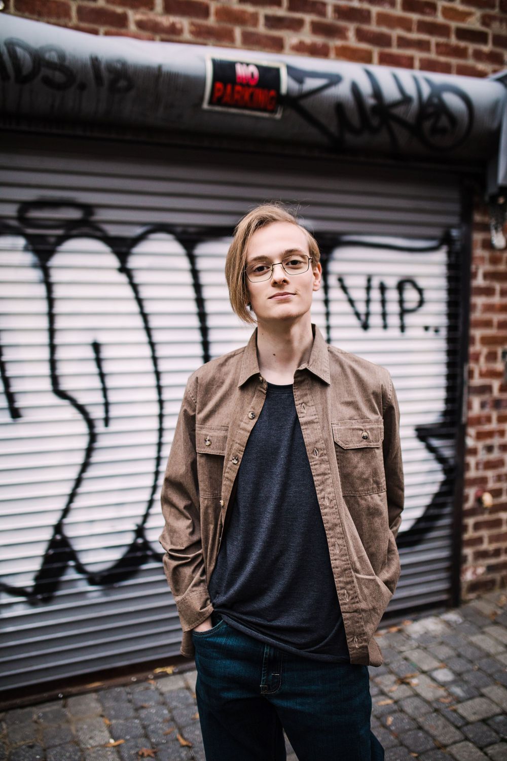 Young person in brown jacket standing before graffiti-covered garage door in urban setting.