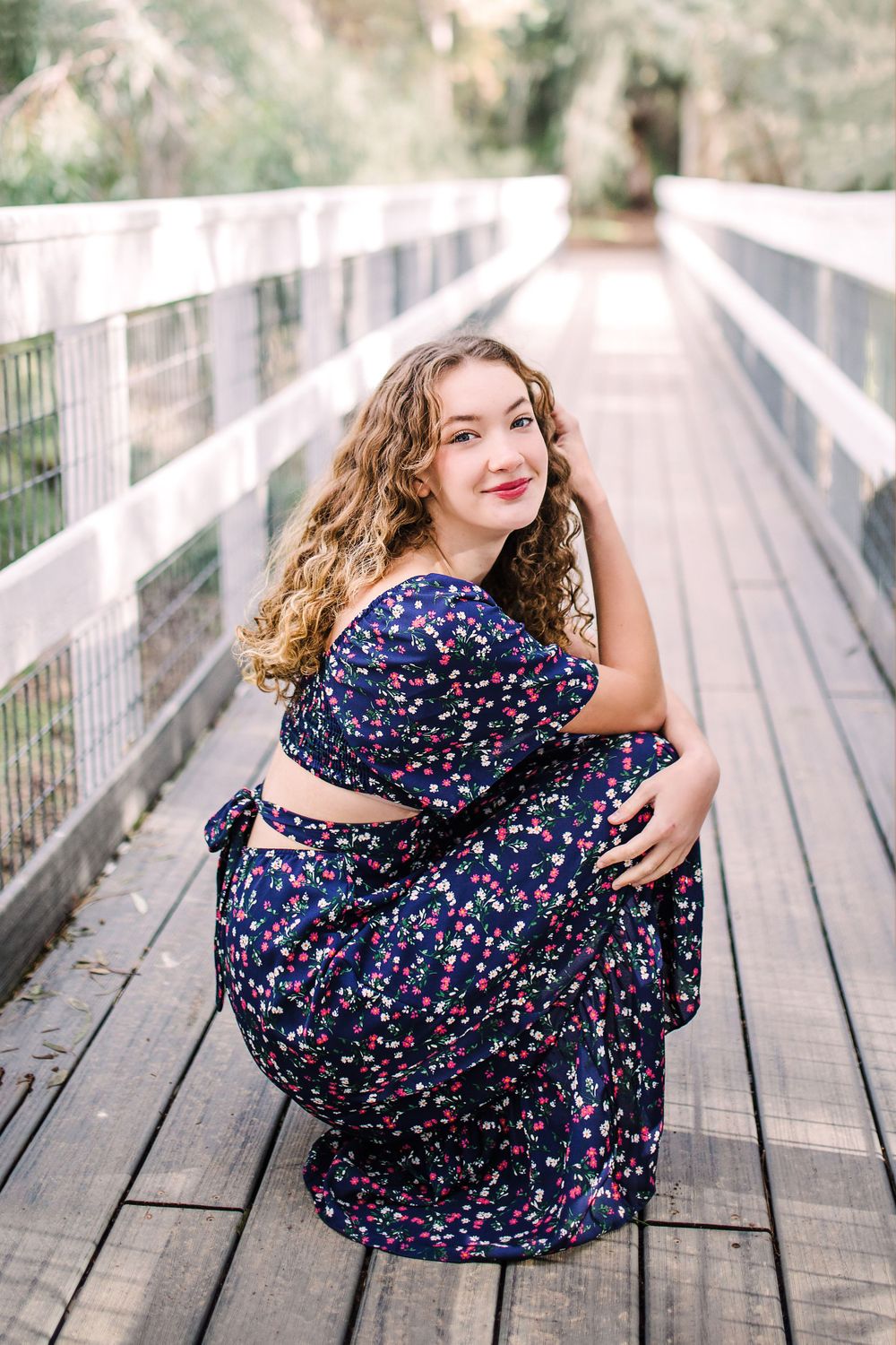 Young woman in navy floral dress crouching on a wooden bridge smiling at camera.