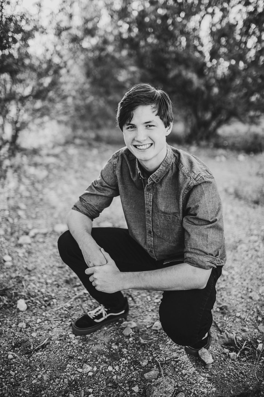 Black and white portrait of smiling teen boy crouching on gravel path outdoors.
