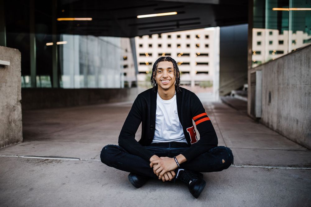 Laughing young man with braids in black cardigan sits cross-legged on ground under urban structure.