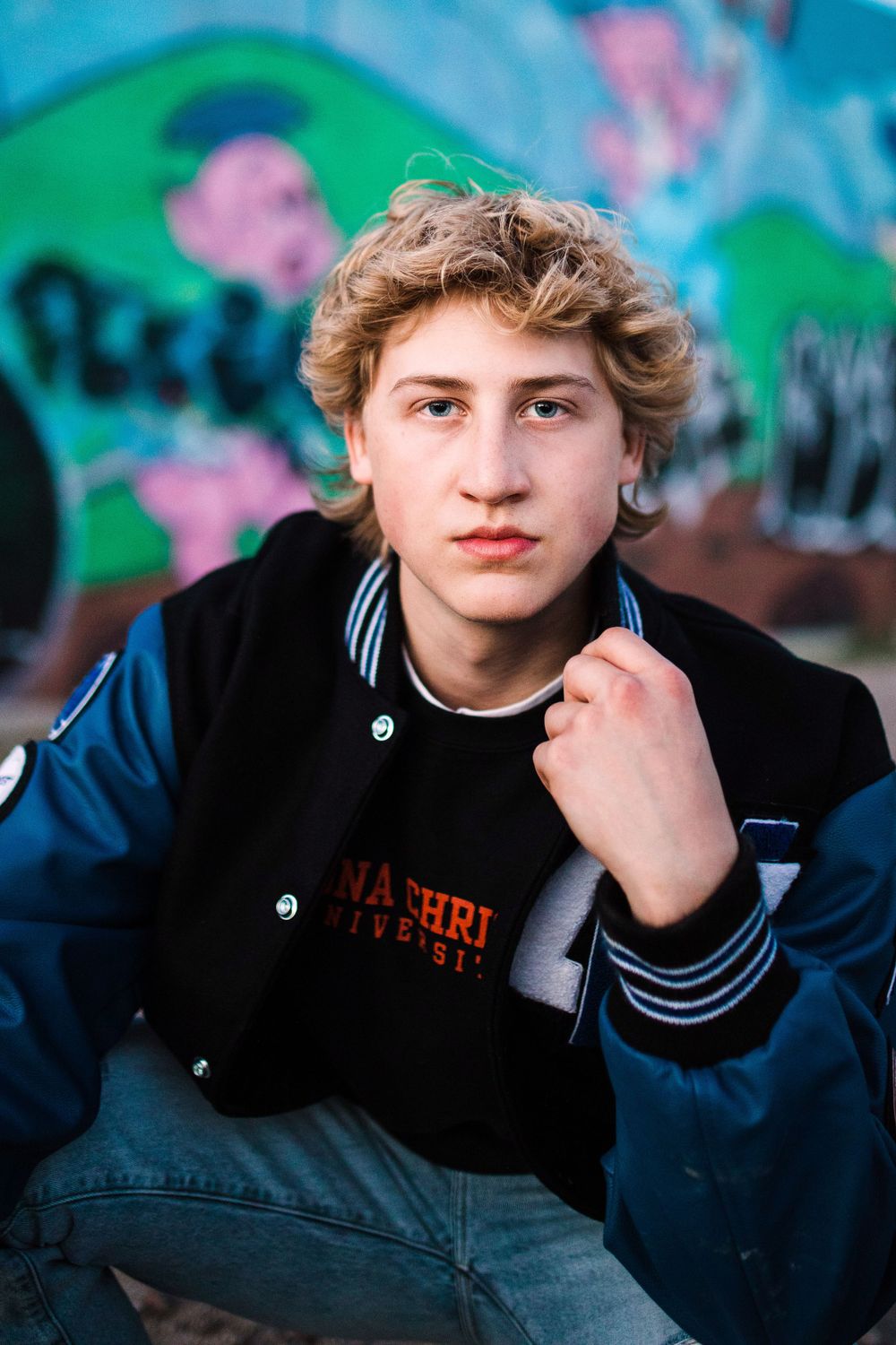 Teen boy in varsity jacket posing confidently in front of colorful graffiti wall.