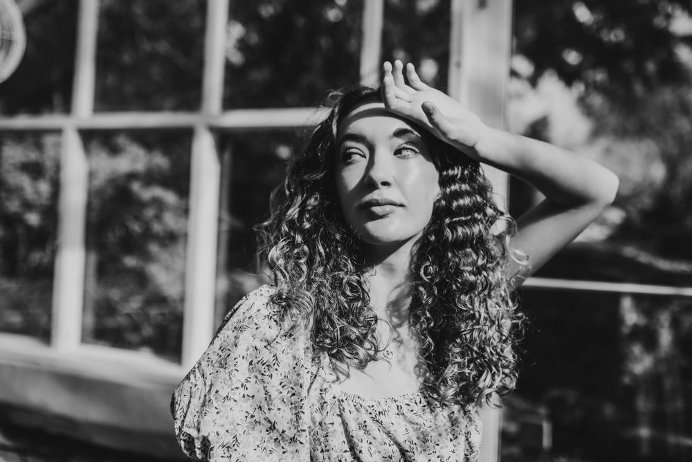 Black and white portrait of curly-haired woman shielding eyes from sunlight outdoors.