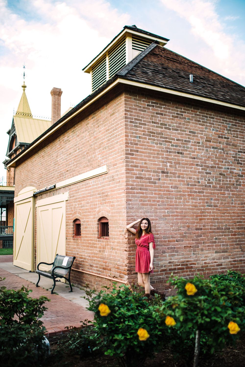 Young woman in red dress posing beside historic brick building with yellow flowers in foreground.