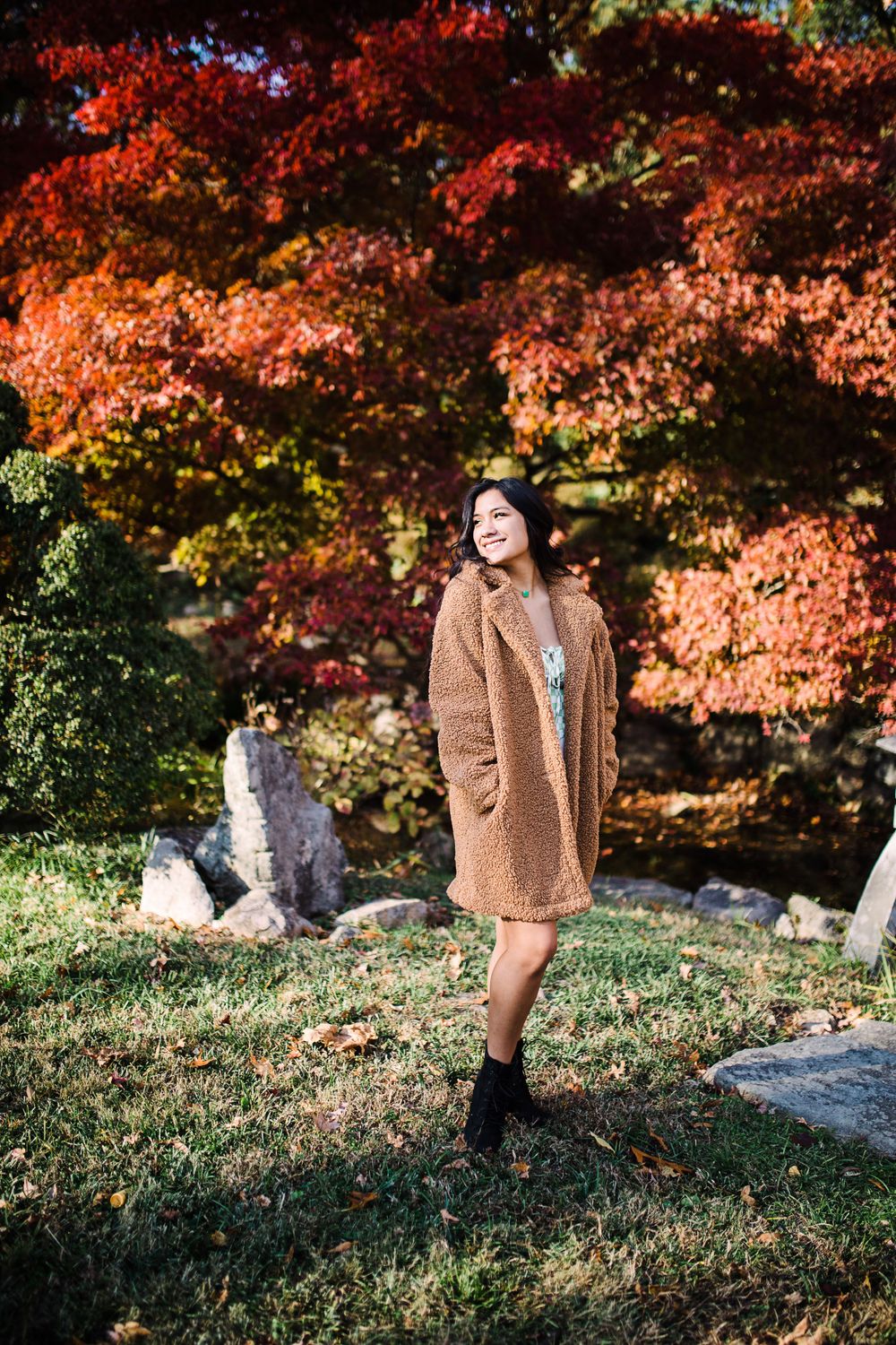 Smiling teen girl in tan teddy coat standing among vibrant red autumn foliage in park.