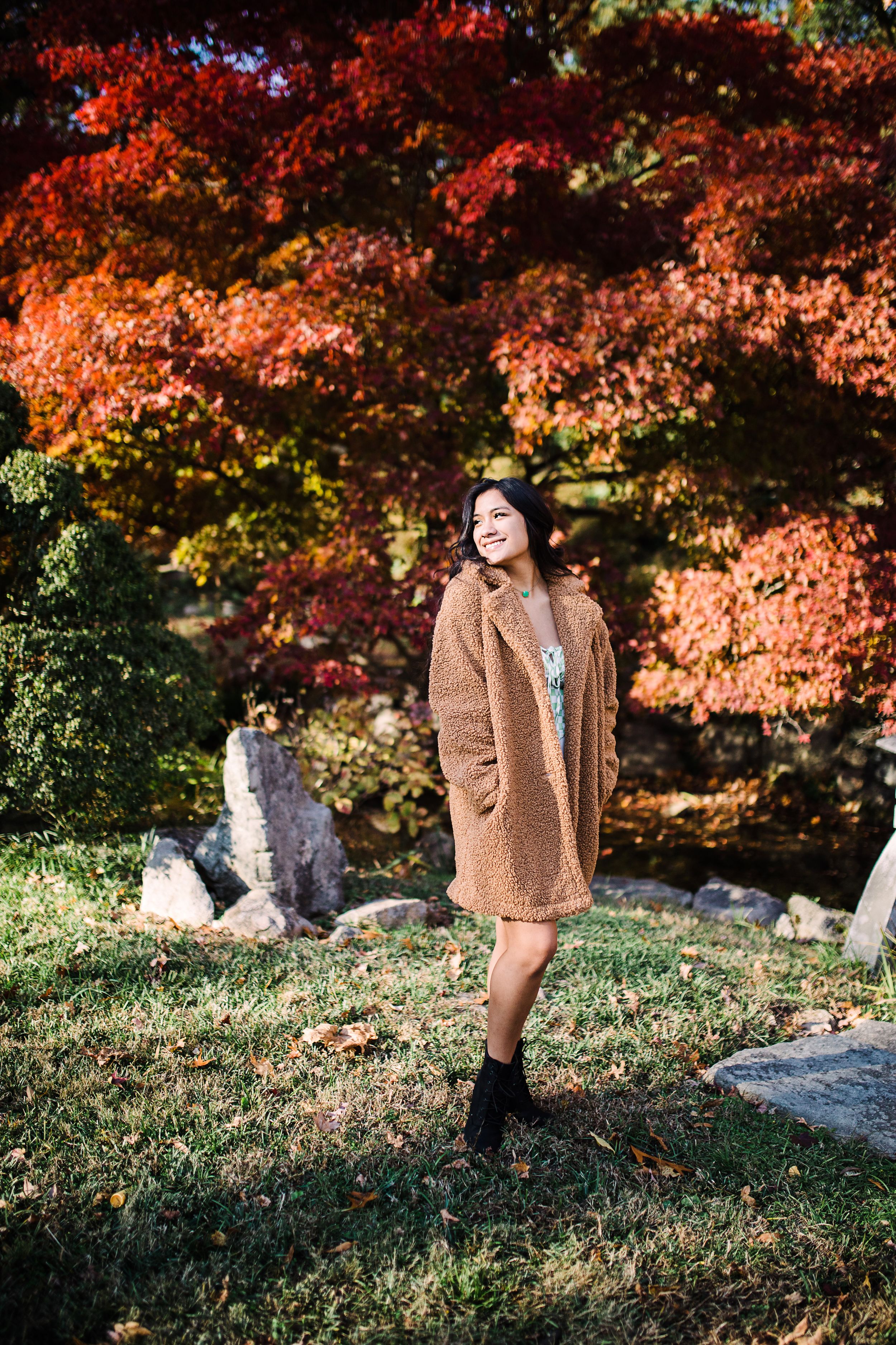 Smiling teen girl in tan teddy coat standing among vibrant red autumn foliage in park.