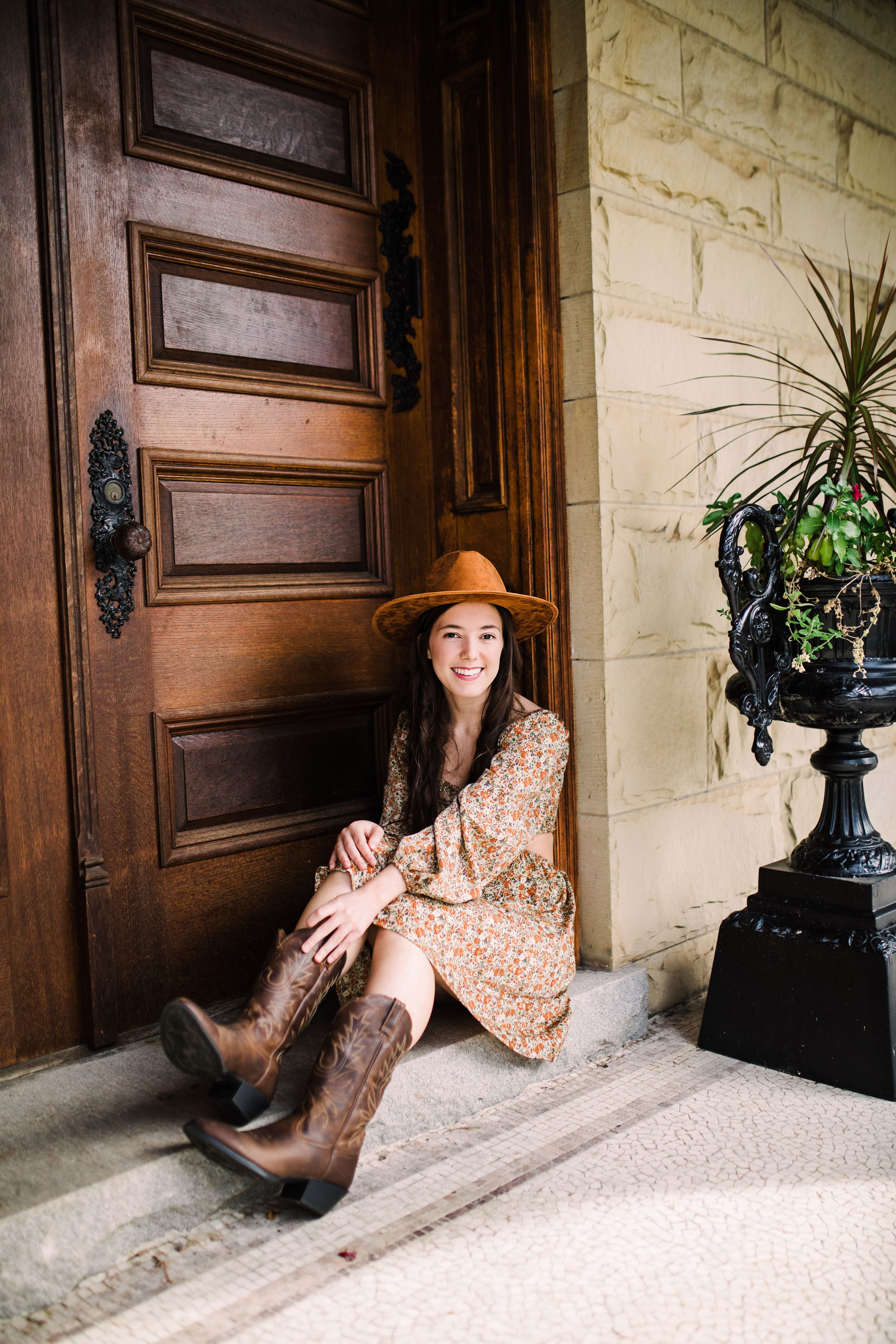 Smiling young woman in floral dress and hat sitting on steps beside ornate wooden door.