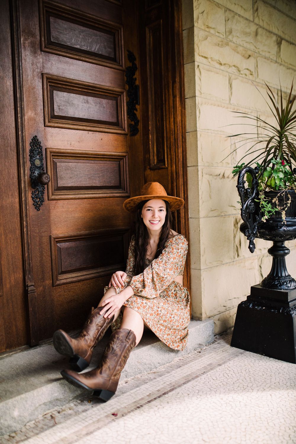 Smiling young woman in floral dress and hat sitting on steps beside ornate wooden door.