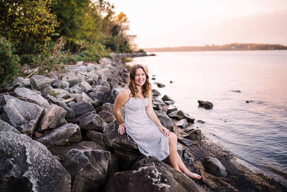 Cheerful young woman in white dress sitting on rocky shore beside calm water during golden sunset.