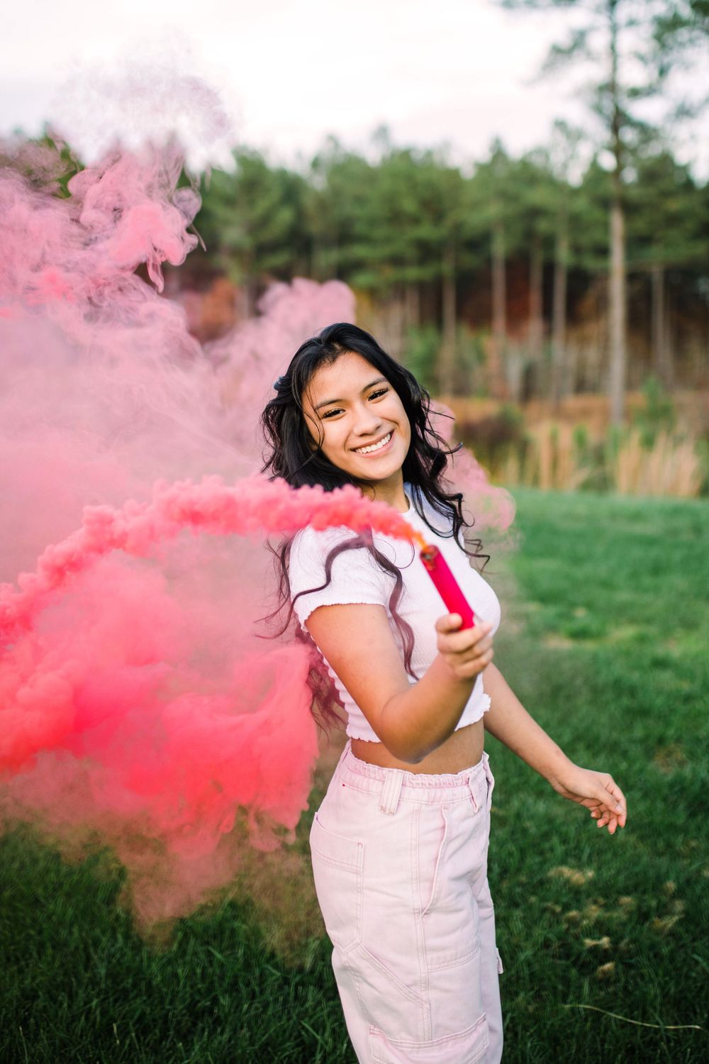Young woman in white crop top smiling while holding a pink smoke bomb in a green field.