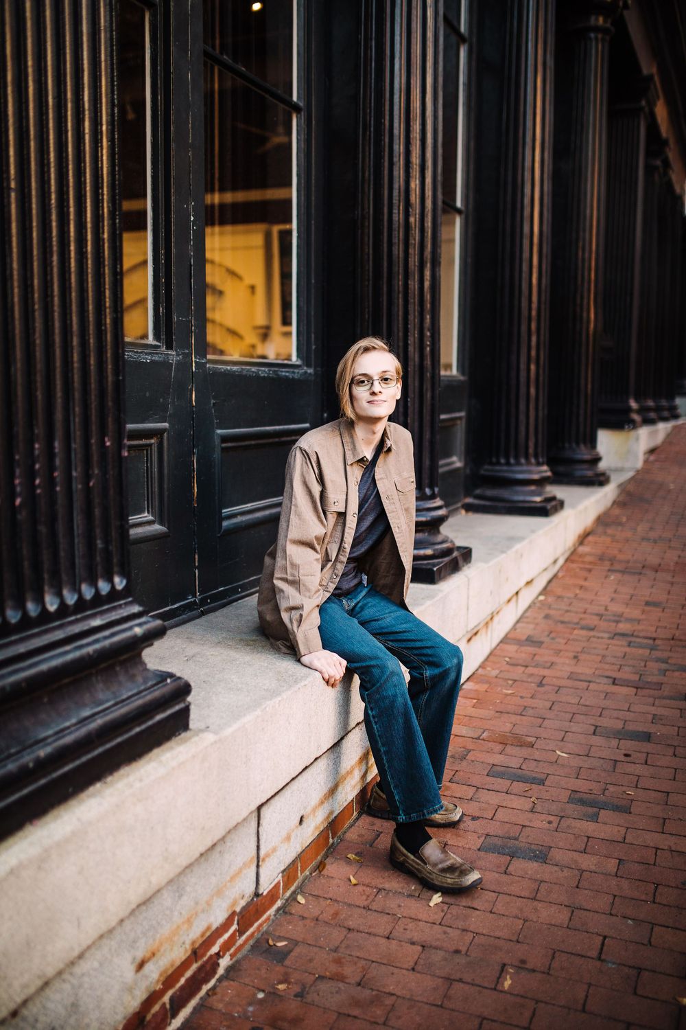 Young man in beige jacket and teal pants sitting on ledge outside historic brick building.