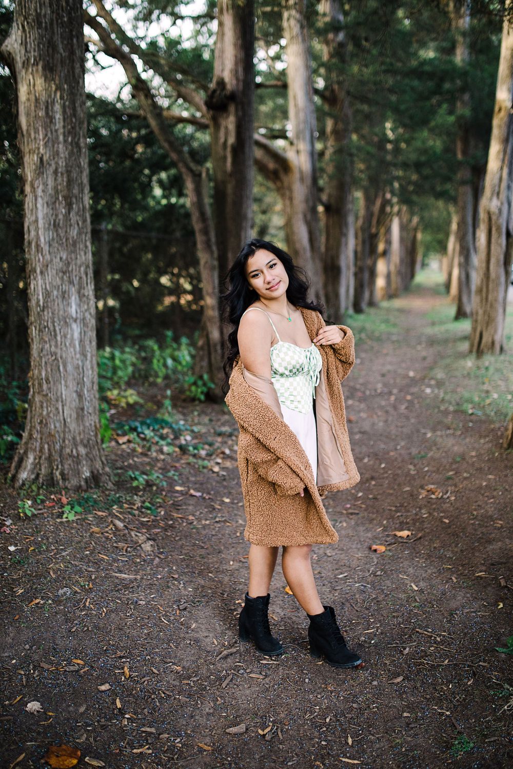 Young woman in brown cardigan and white dress poses on a serene tree-lined forest path.
