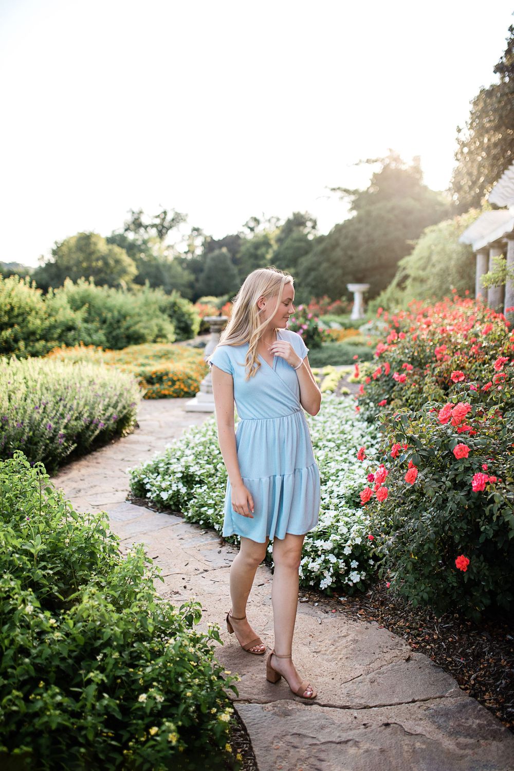 Blonde teen in light blue dress walking through colorful rose garden in golden sunlight.