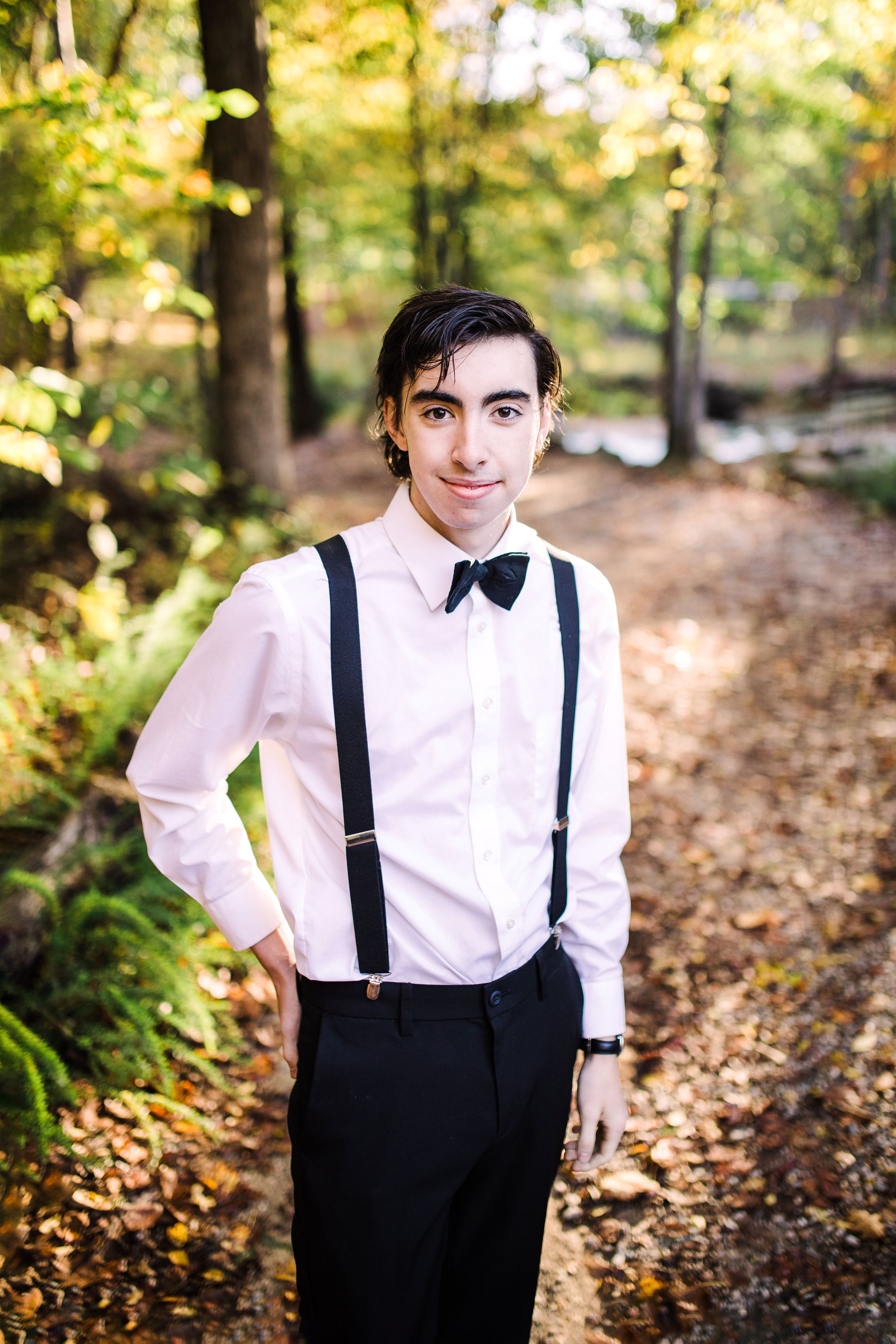 Young man in white dress shirt, black suspenders and bow tie smiling on leaf-covered forest path.