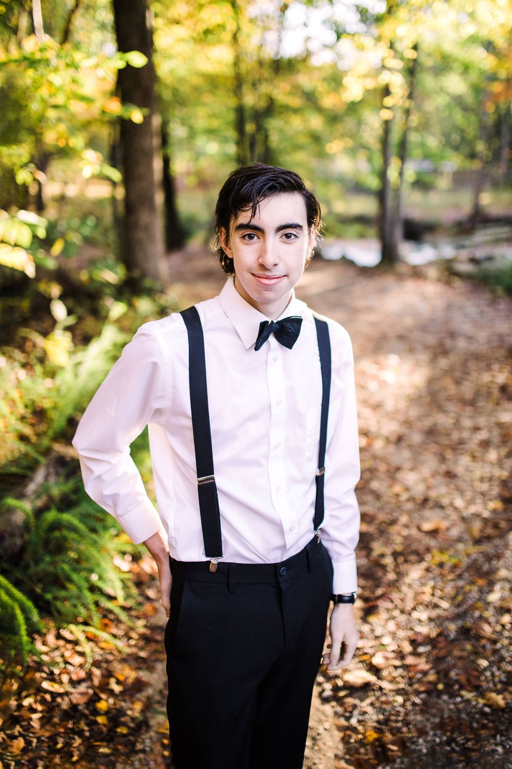 Young man in white dress shirt, black suspenders and bow tie smiling on leaf-covered forest path.