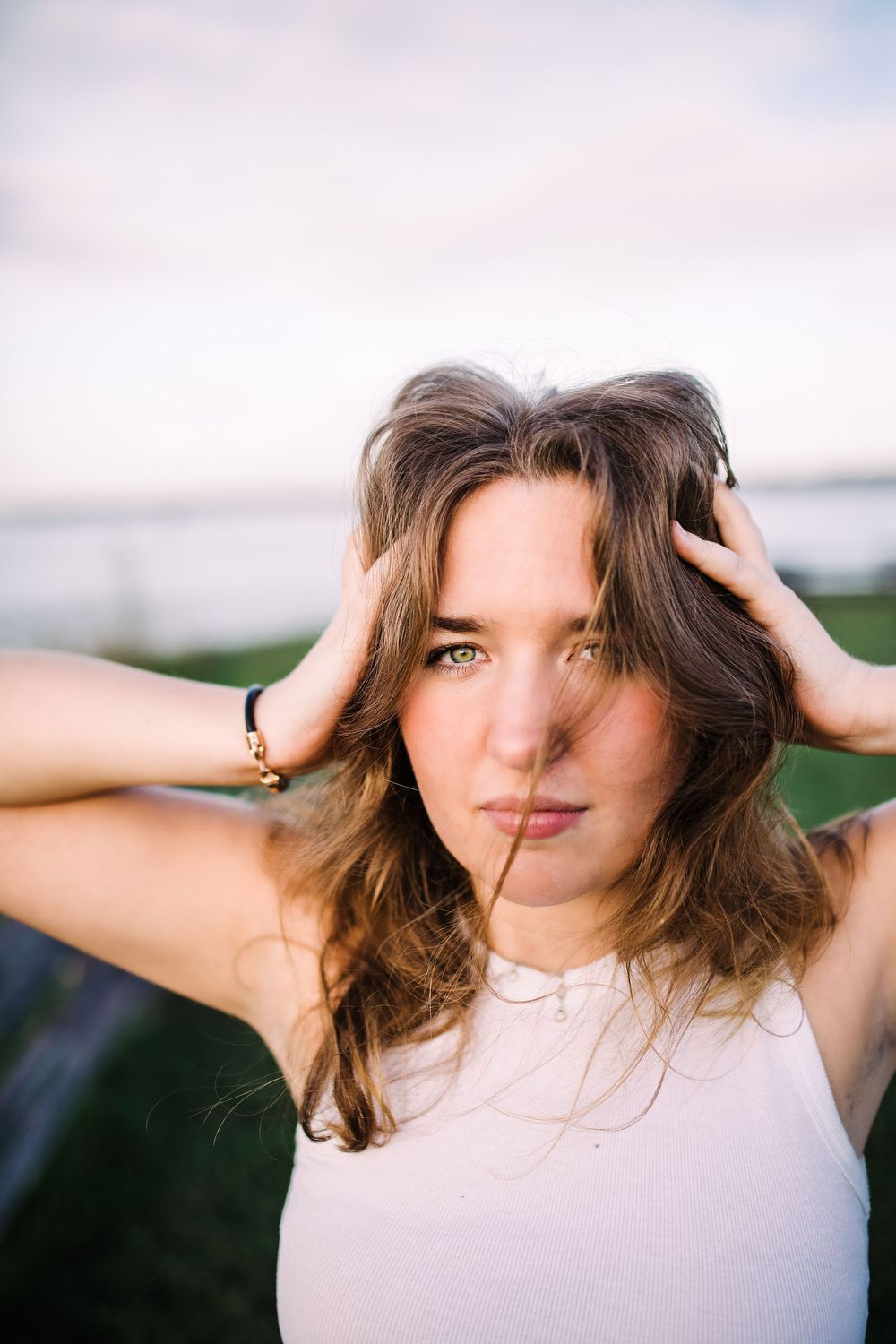 Young woman with green eyes and wavy hair poses outdoors near water in white tank top.