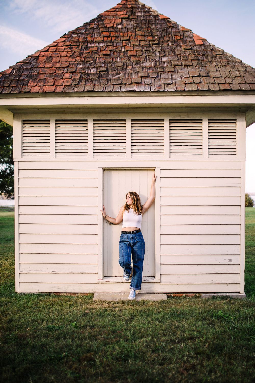 Young woman in white top and jeans leaning against a white wooden outbuilding with brick roof.