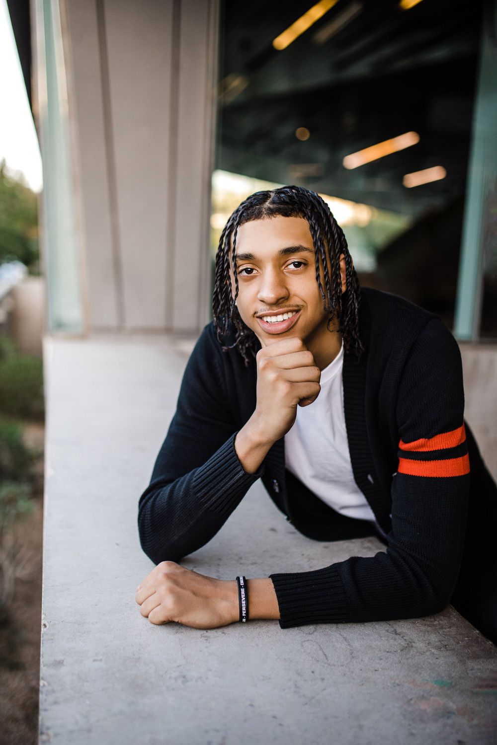 Smiling young man with braids in black cardigan rests chin on hand while leaning on ledge.