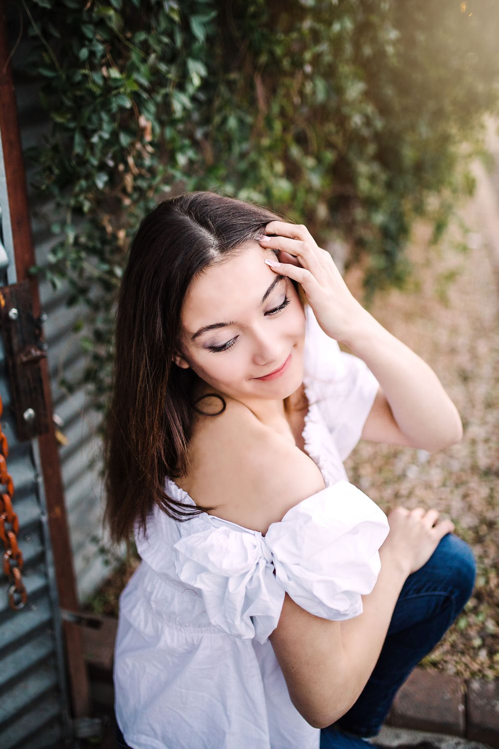 Beautiful brunette woman in white off-shoulder top posing near ivy-covered wall outdoors.