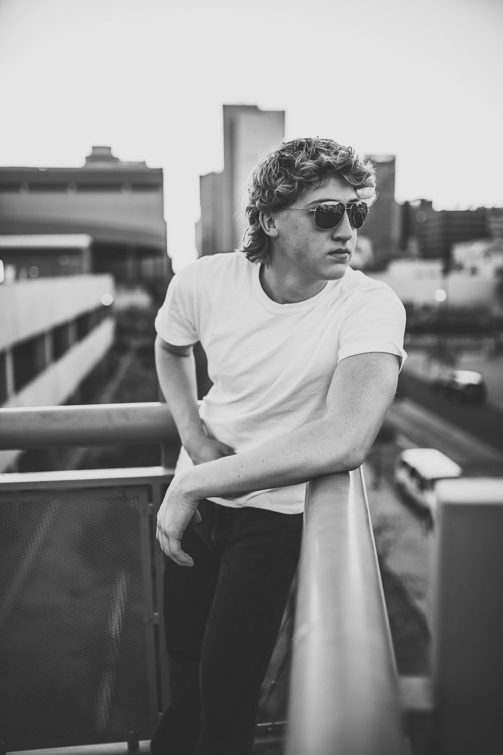 Young man in white tee and sunglasses leaning on railing with city skyline behind.