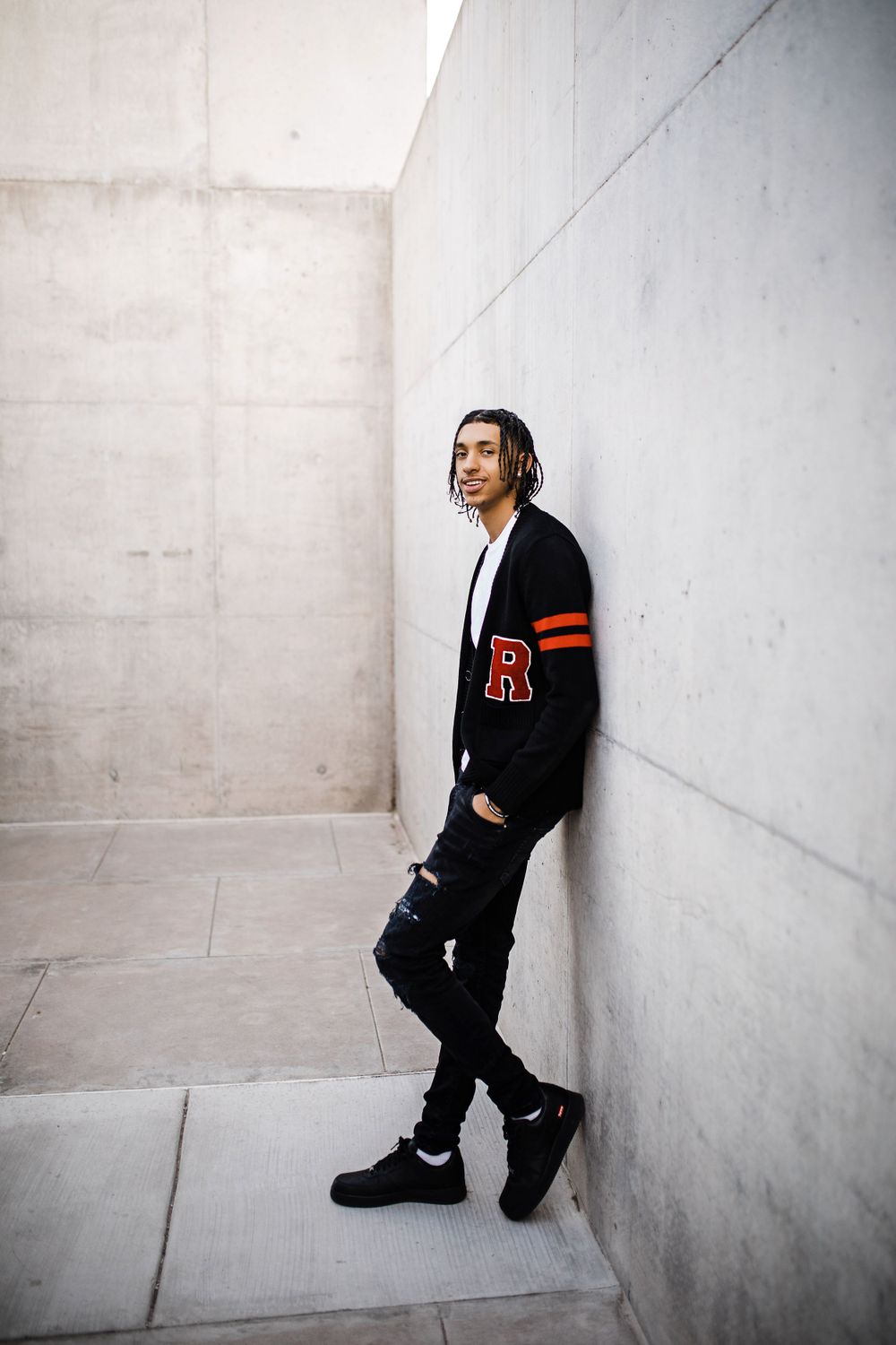 Young man in black varsity cardigan with R letter patch leans against concrete wall outdoors.