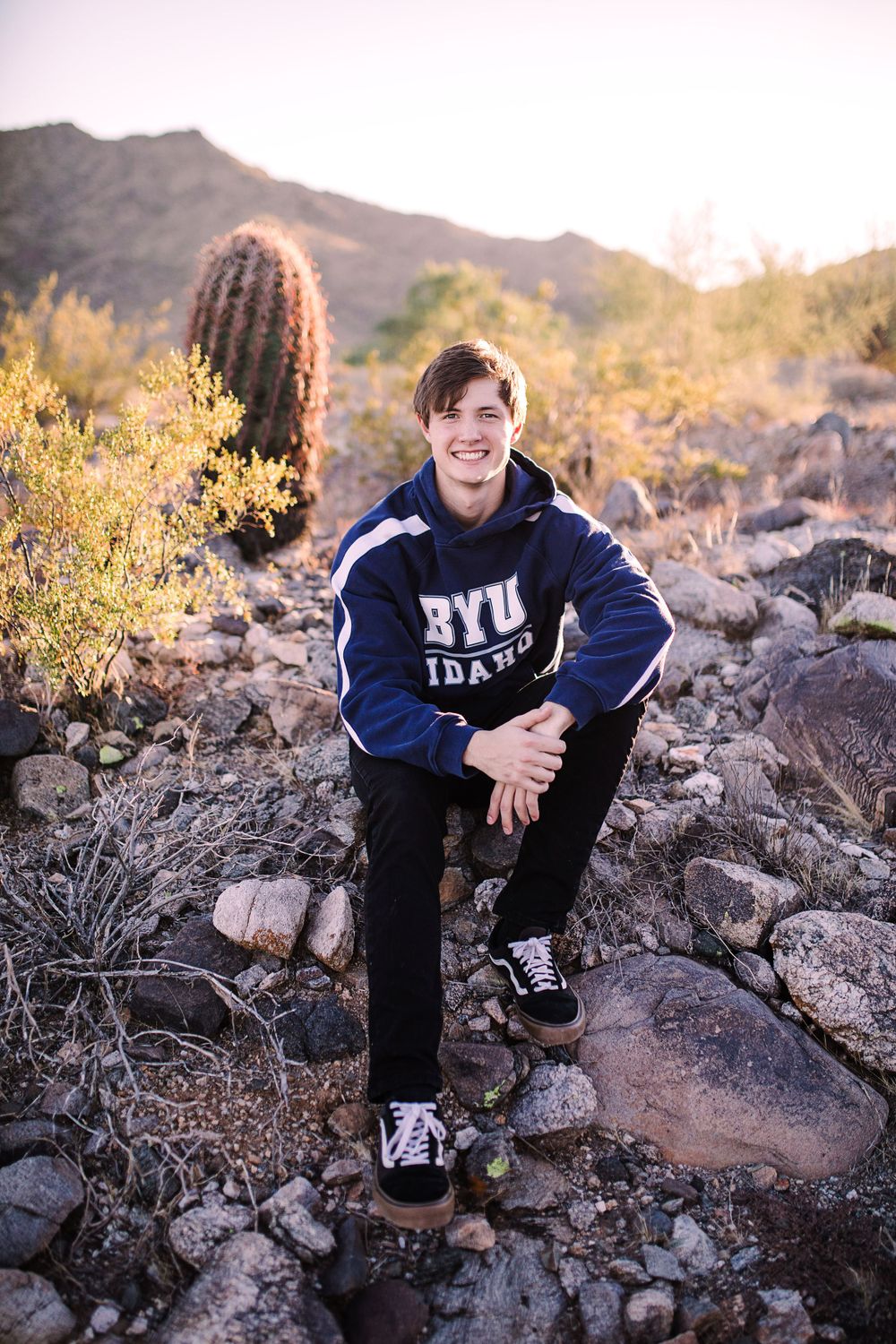 Young man in BYU Idaho hoodie sitting on rocks in desert landscape with cactus and mountains at sunset.