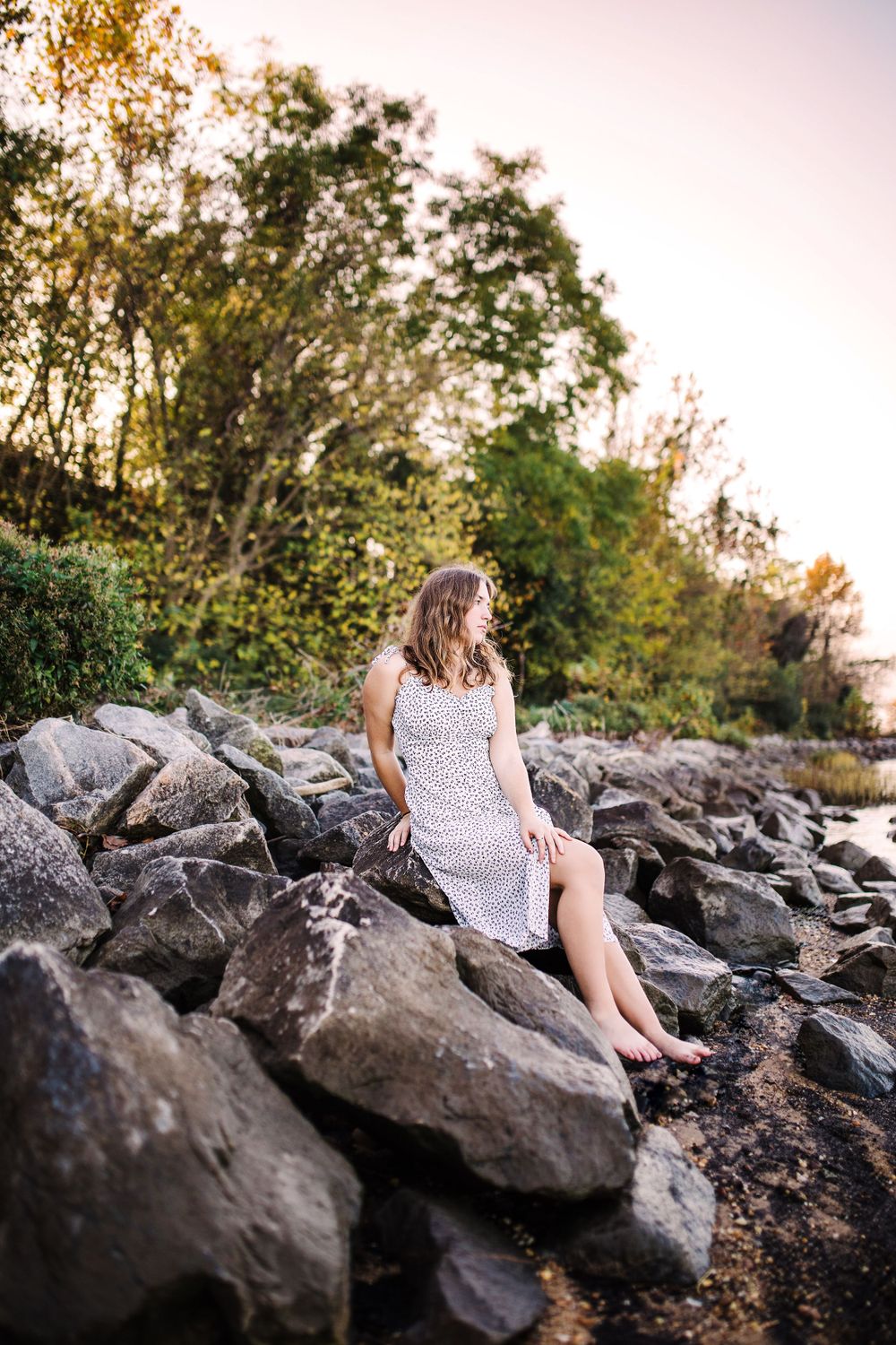 Young woman in white floral dress sitting barefoot on large rocks by a serene waterfront at dusk.