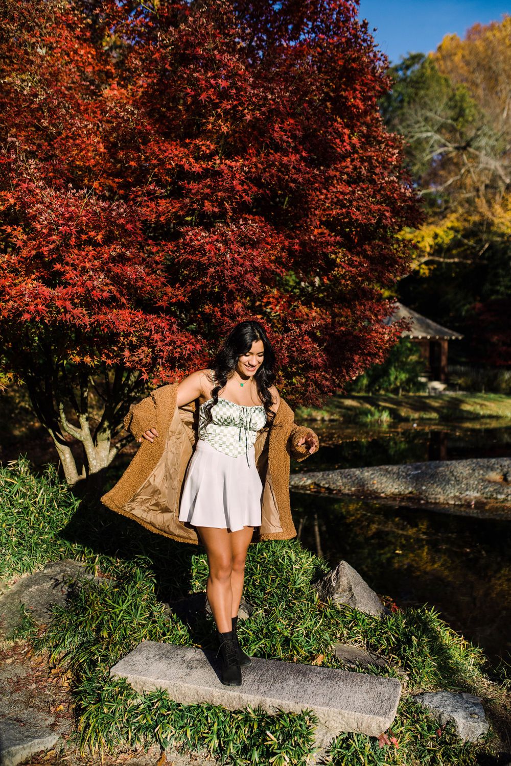 Young woman in white dress opens tan coat while standing on rocks near vibrant red autumn trees.