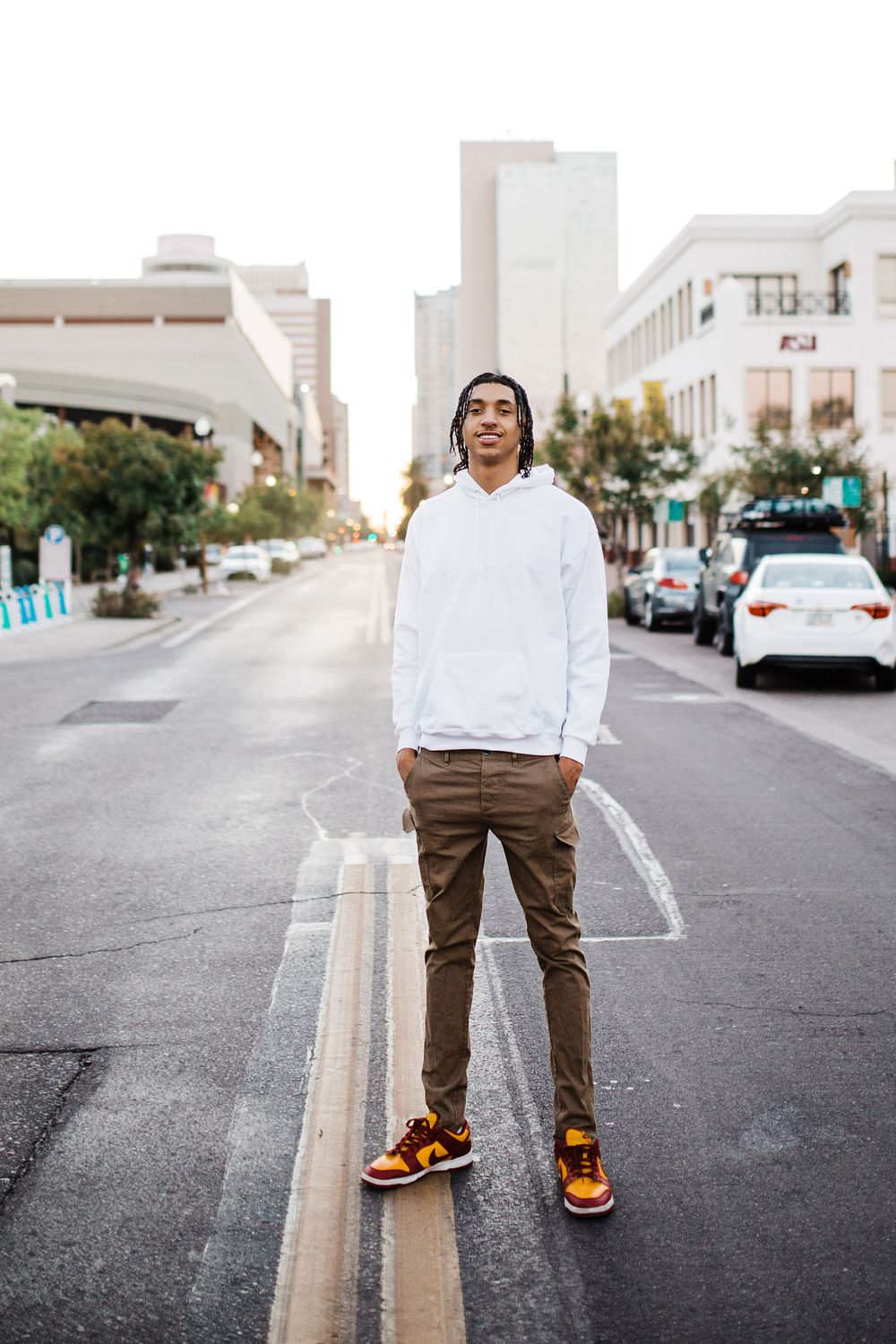 Young man in white hoodie and brown pants standing on urban street with city buildings behind him.