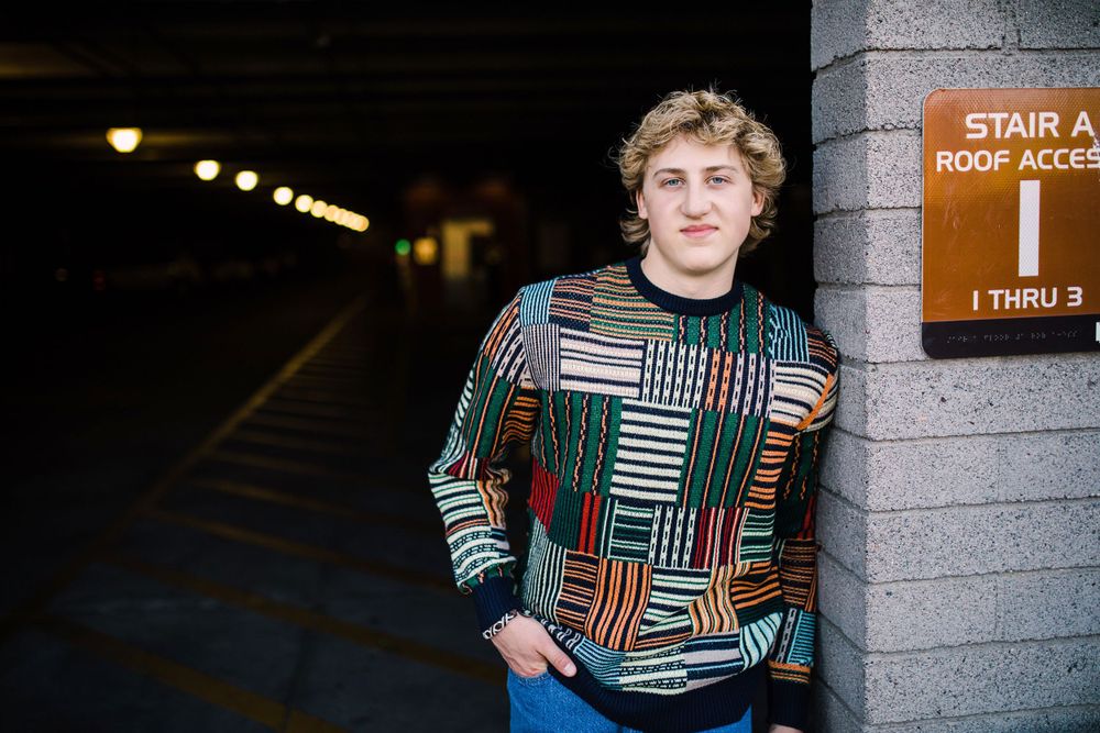 Young man in colorful geometric sweater leaning against wall near stair access sign in parking garage.