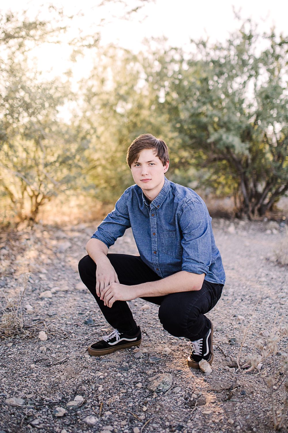 Teen boy in denim shirt crouching on dirt path surrounded by desert shrubs.