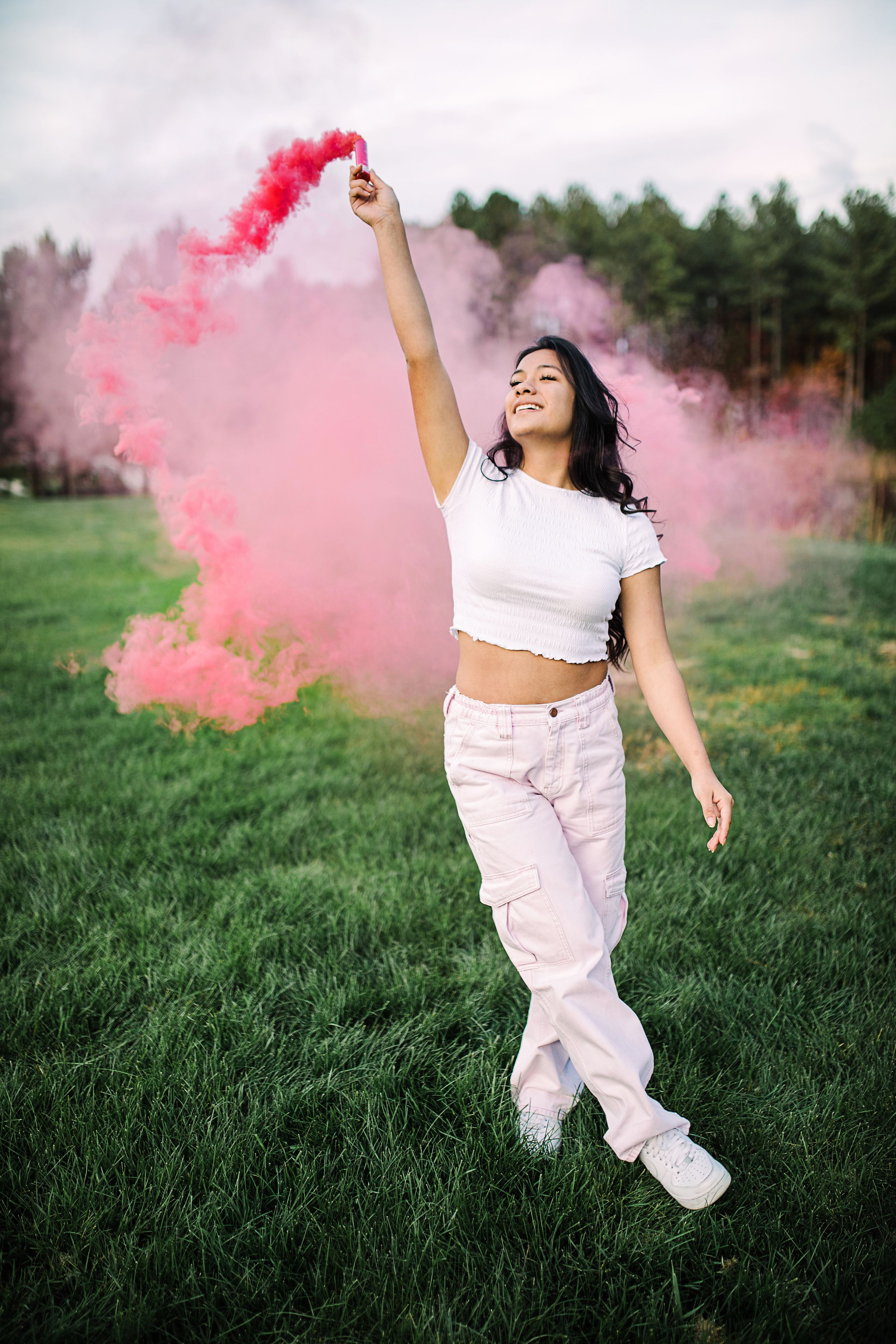 Young woman in white outfit raising a pink smoke bomb overhead while walking through a green field.