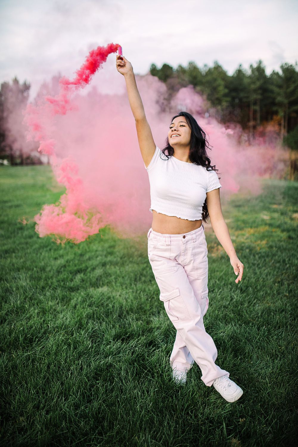 Young woman in white outfit raising a pink smoke bomb overhead while walking through a green field.