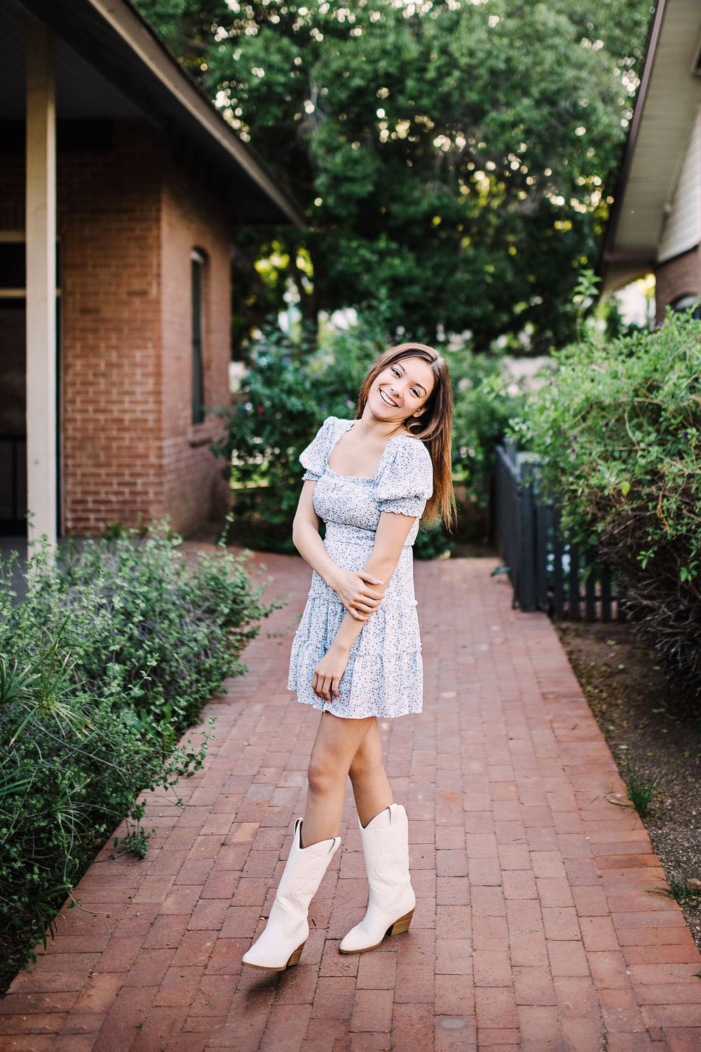 Smiling young woman in floral dress and white cowboy boots on a brick garden path.