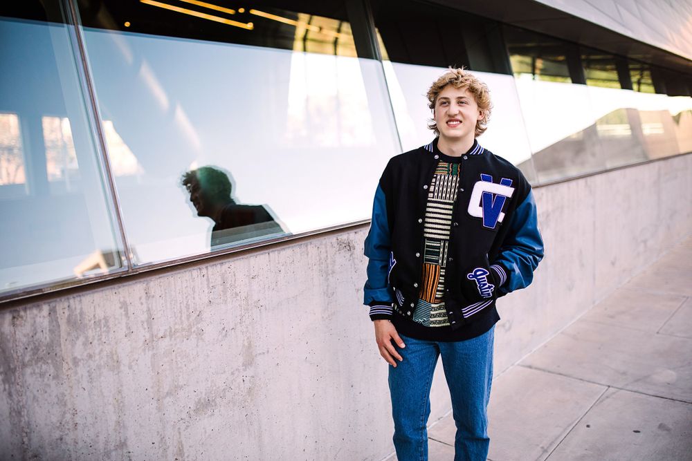 Teen boy in blue varsity jacket standing by glass building reflection outdoors.
