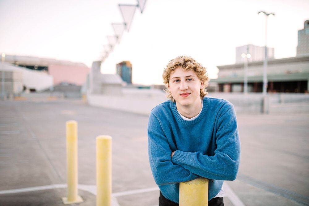 Smiling teen leaning on yellow bollard in open parking lot at golden hour.