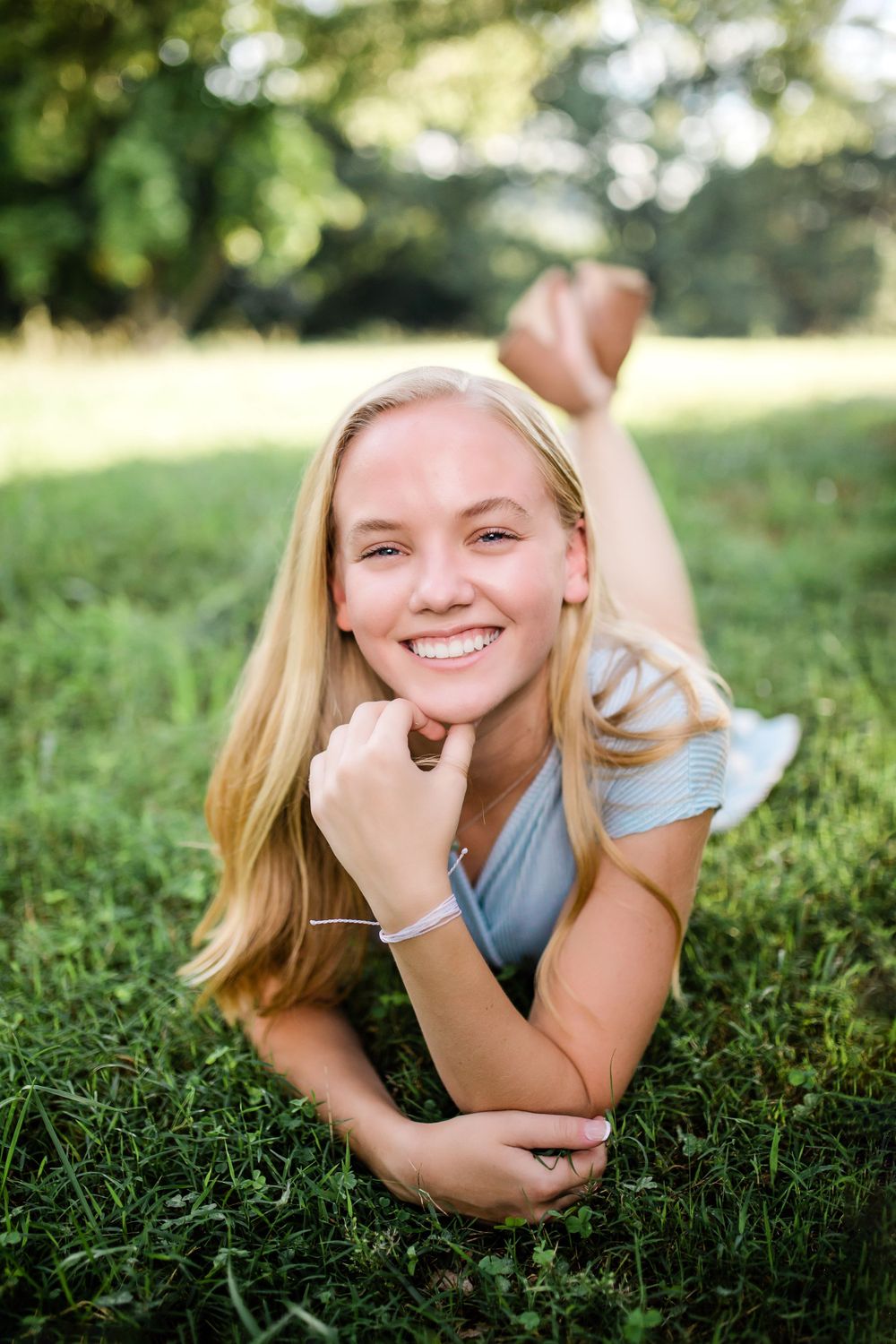 Cheerful blonde teen girl lying on green grass smiling brightly in light blue top outdoors.