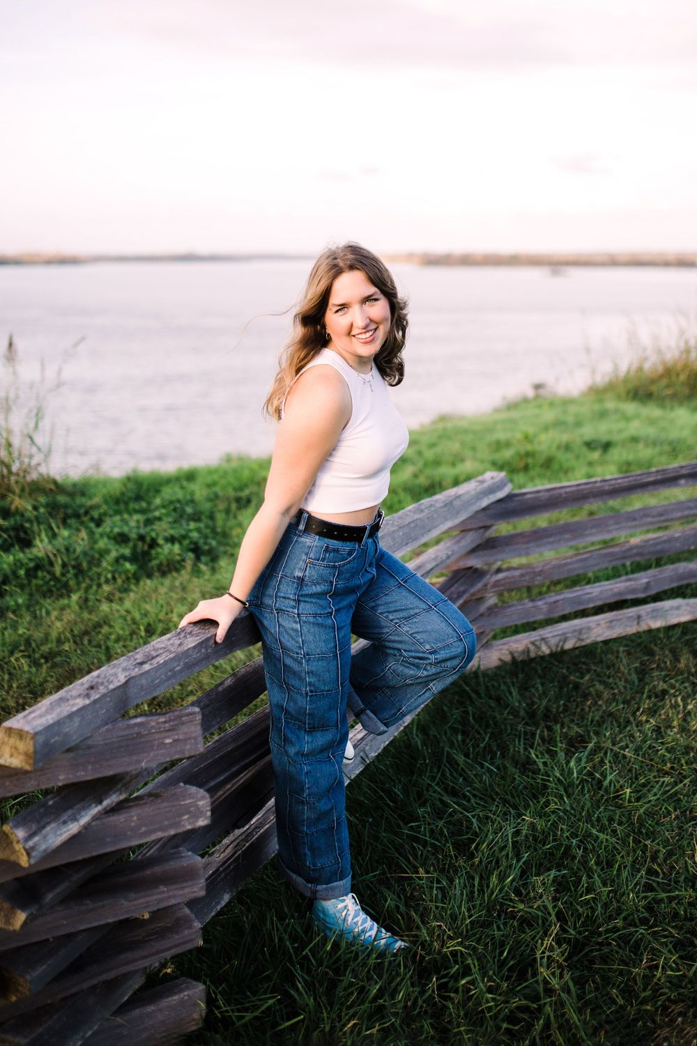 Smiling young woman in white crop top and jeans leans on wooden fence near water.