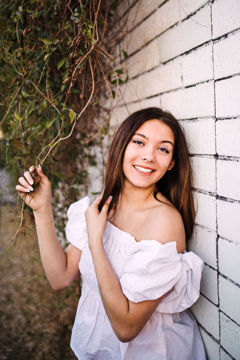 Smiling young woman in white off-shoulder dress leaning against white brick wall with ivy vines.