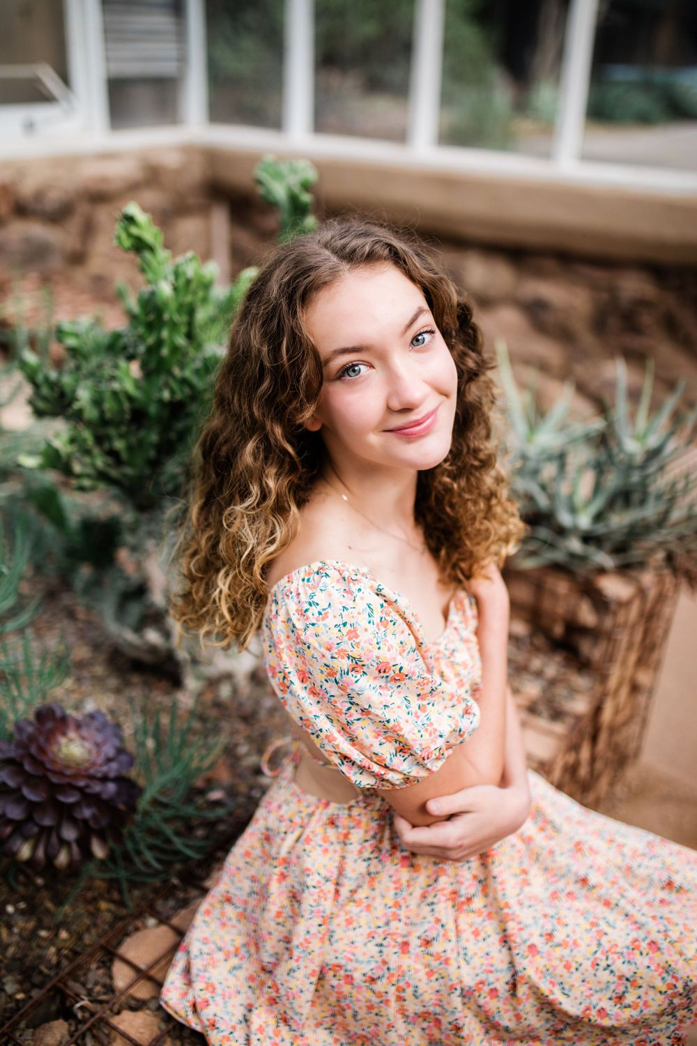 Smiling young woman in off-shoulder floral dress sitting among succulents in greenhouse garden.
