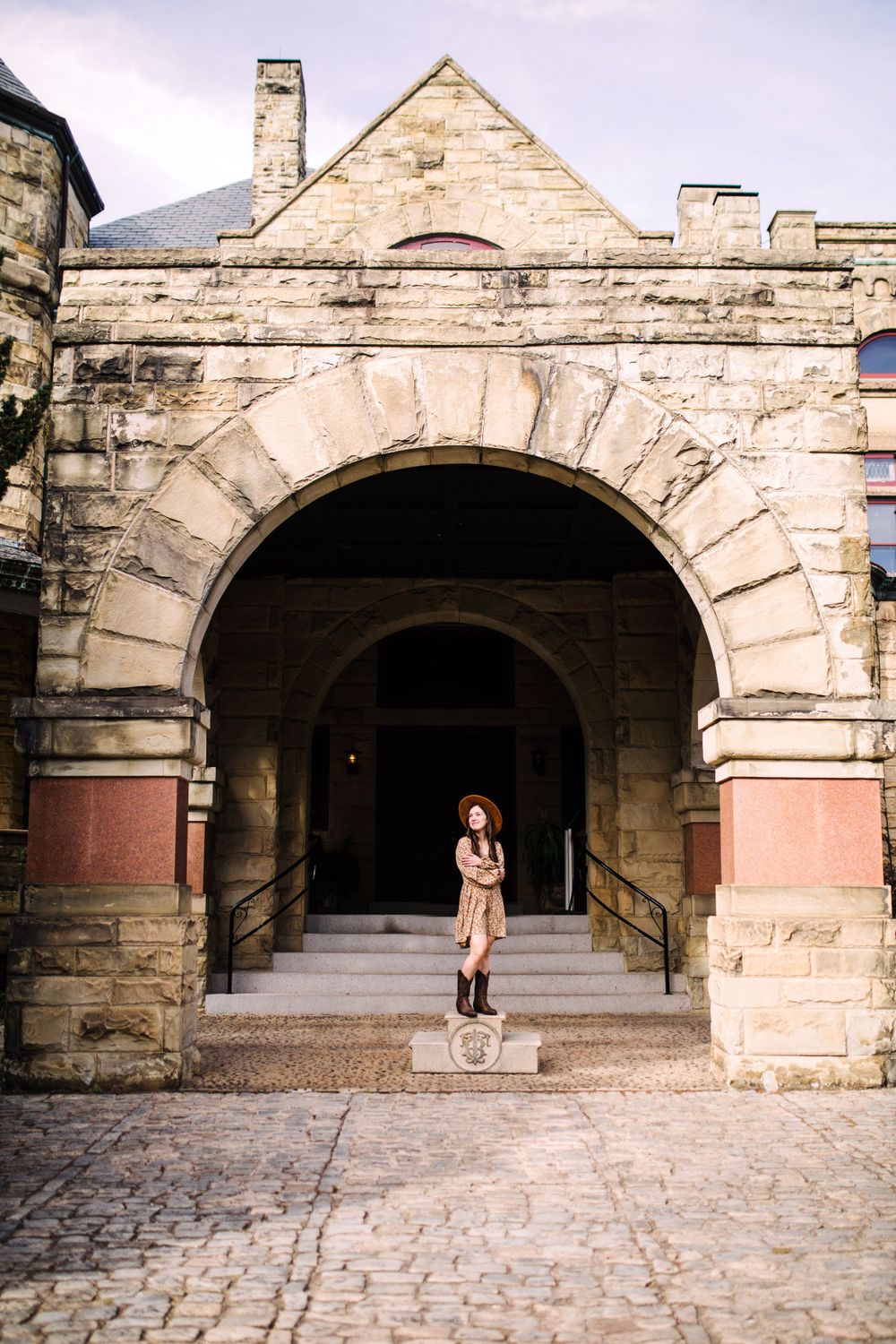 Young woman standing under large stone arch entrance of historic building on cobblestone plaza.