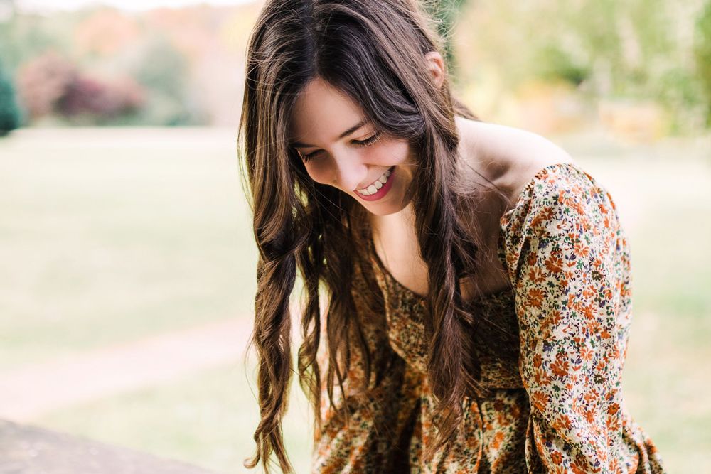 Young woman with long dark hair smiling downward wearing floral dress outdoors in green park setting.