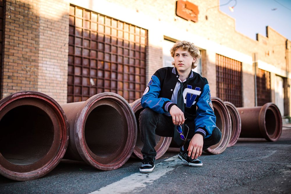 Young man in blue varsity jacket crouching in front of large industrial pipes near brick building.