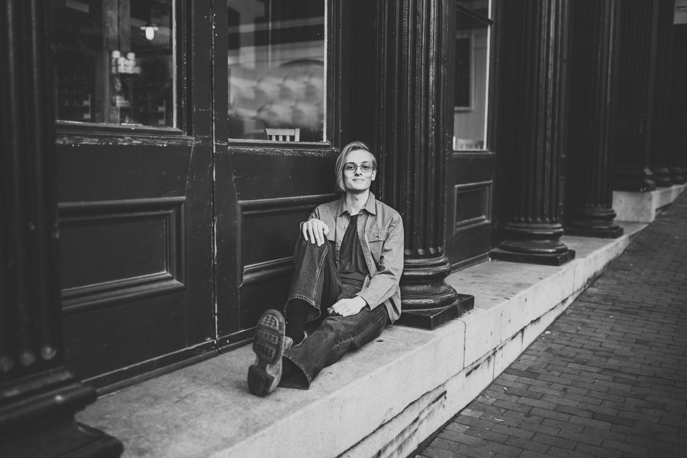 Black and white photo of young man with glasses sitting relaxed on urban storefront steps.