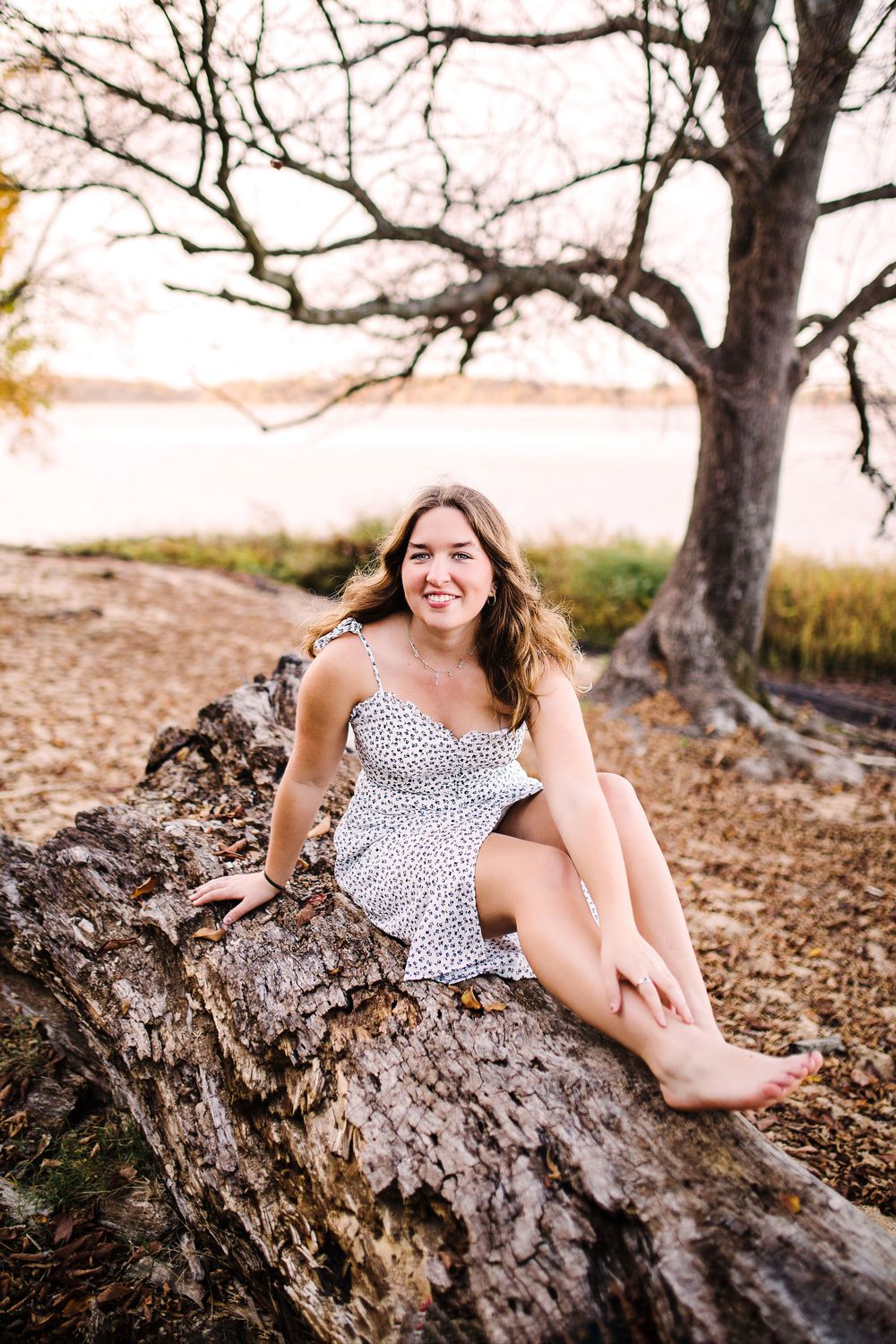 Smiling young woman in white floral sundress sitting on fallen log near lakeside at sunset.