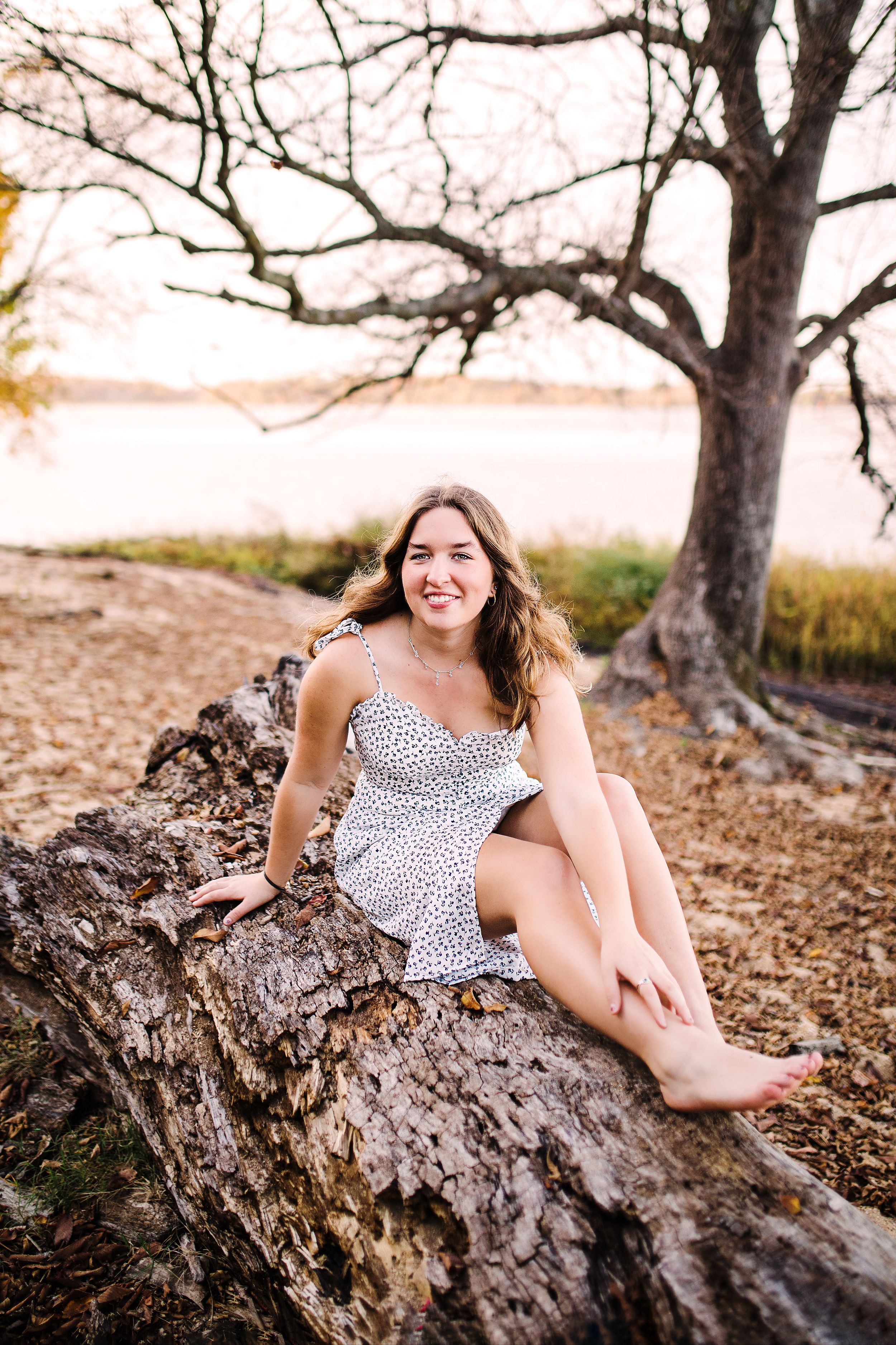 Smiling young woman in white floral sundress sitting on fallen log near lakeside at sunset.