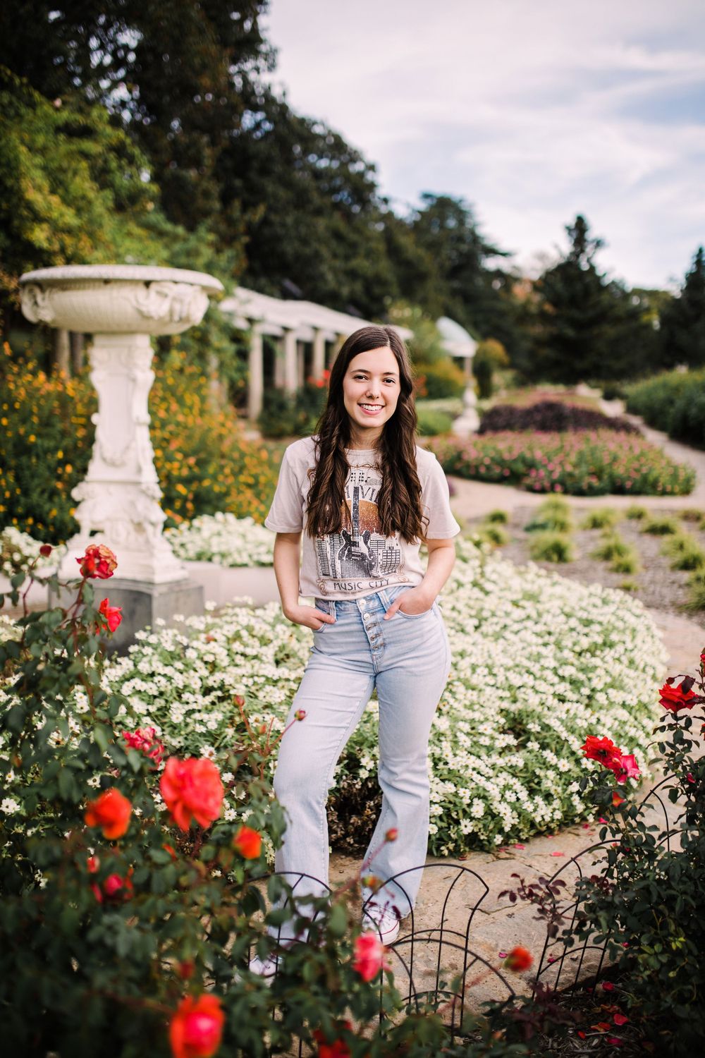 Smiling young woman in Nashville tee and jeans standing among red roses in formal garden.