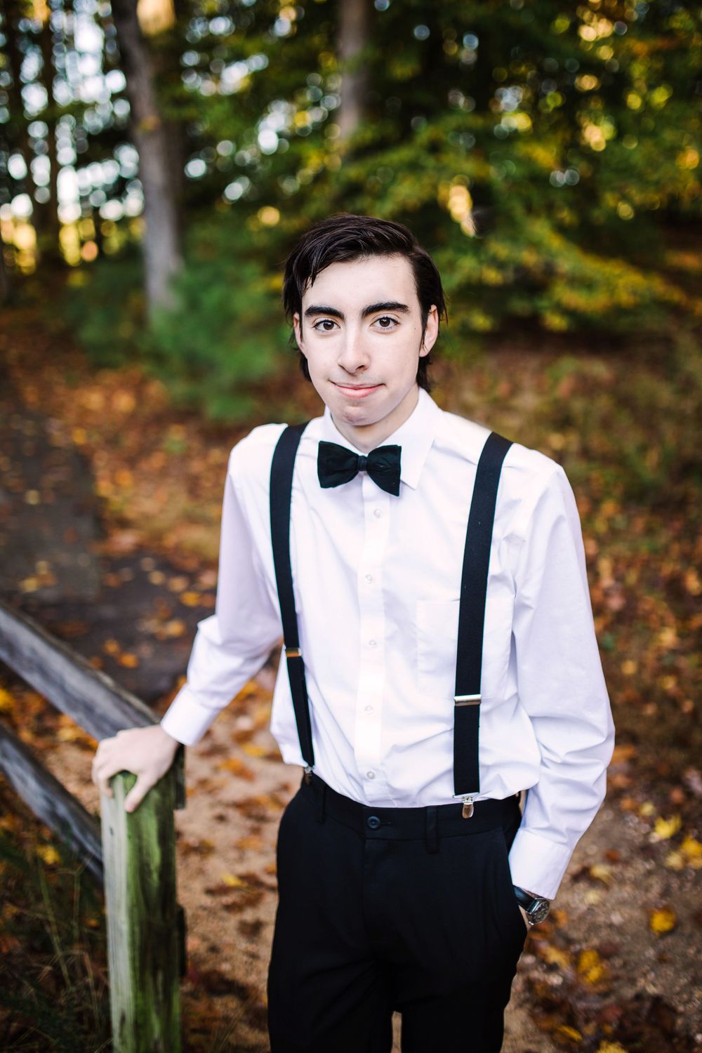 Young man in white shirt, black suspenders and bow tie posing on wooden steps in autumn forest.